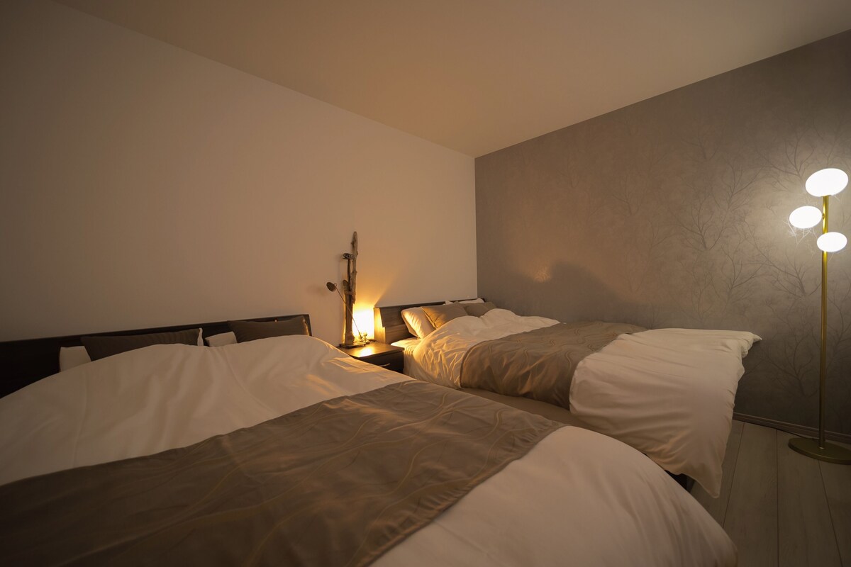A hotel room with two beds, a bedside table with a lamp, and a floor lamp with three lights.
