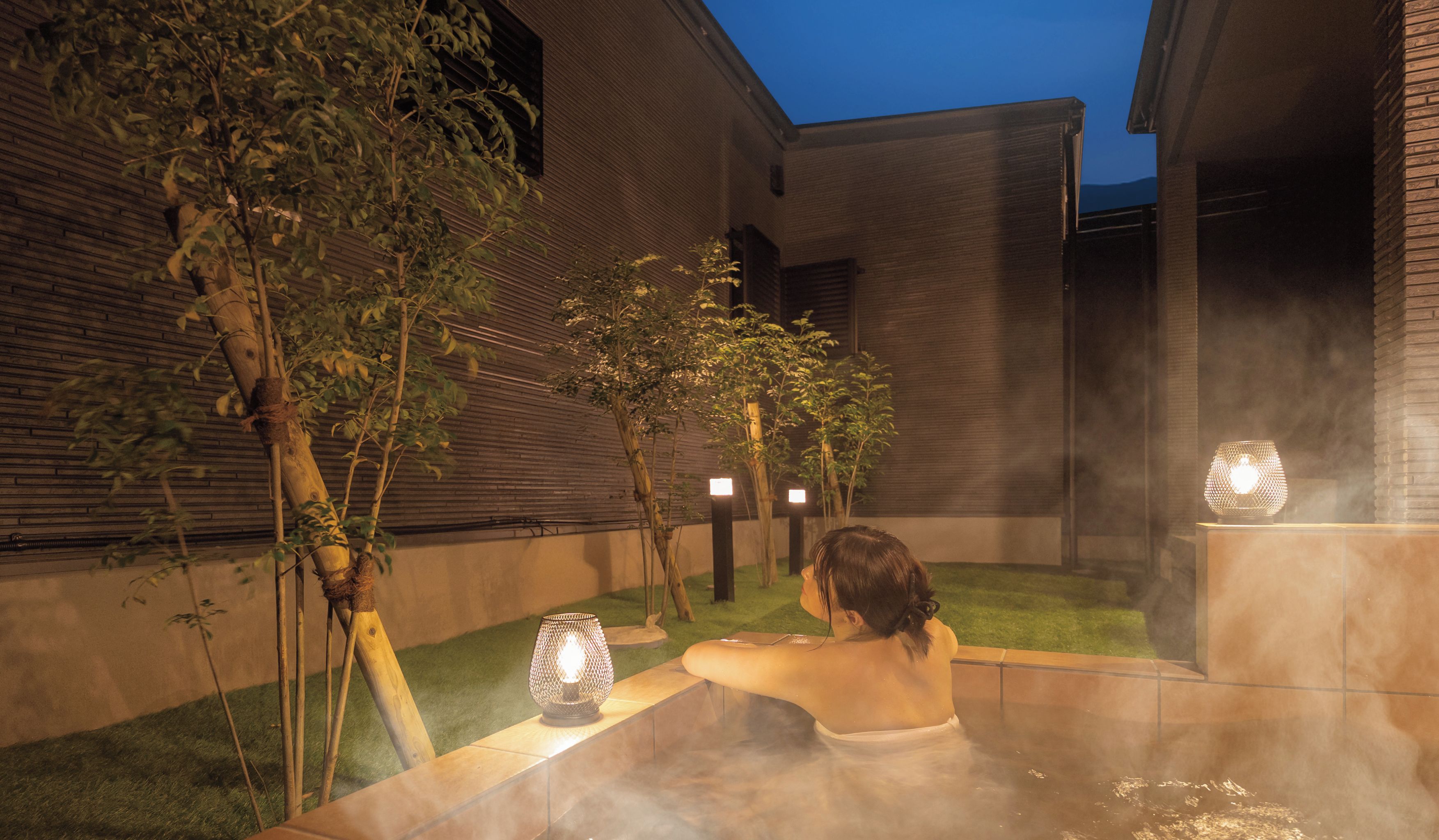 A person relaxing in a hot tub outdoors with a view of a brick building and trees.
