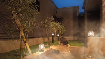 A person relaxing in a hot tub outdoors with a view of a brick building and trees.