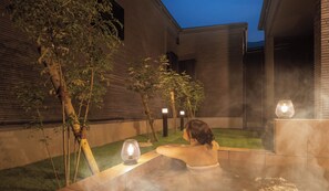 Une personne se détend dans un bain à remous en plein air, avec vue sur un bâtiment en briques et des arbres.