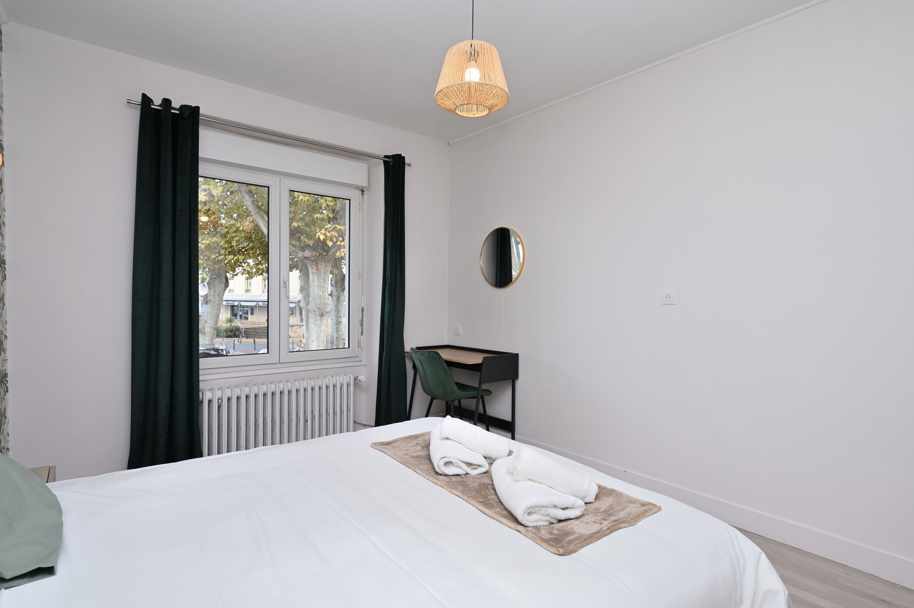 Habitación de hotel con cama, escritorio, silla, ventana con cortinas, radiador y lámpara colgante.