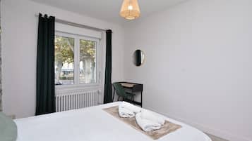 Habitación de hotel con cama, escritorio, silla, ventana con cortinas, radiador y lámpara colgante.