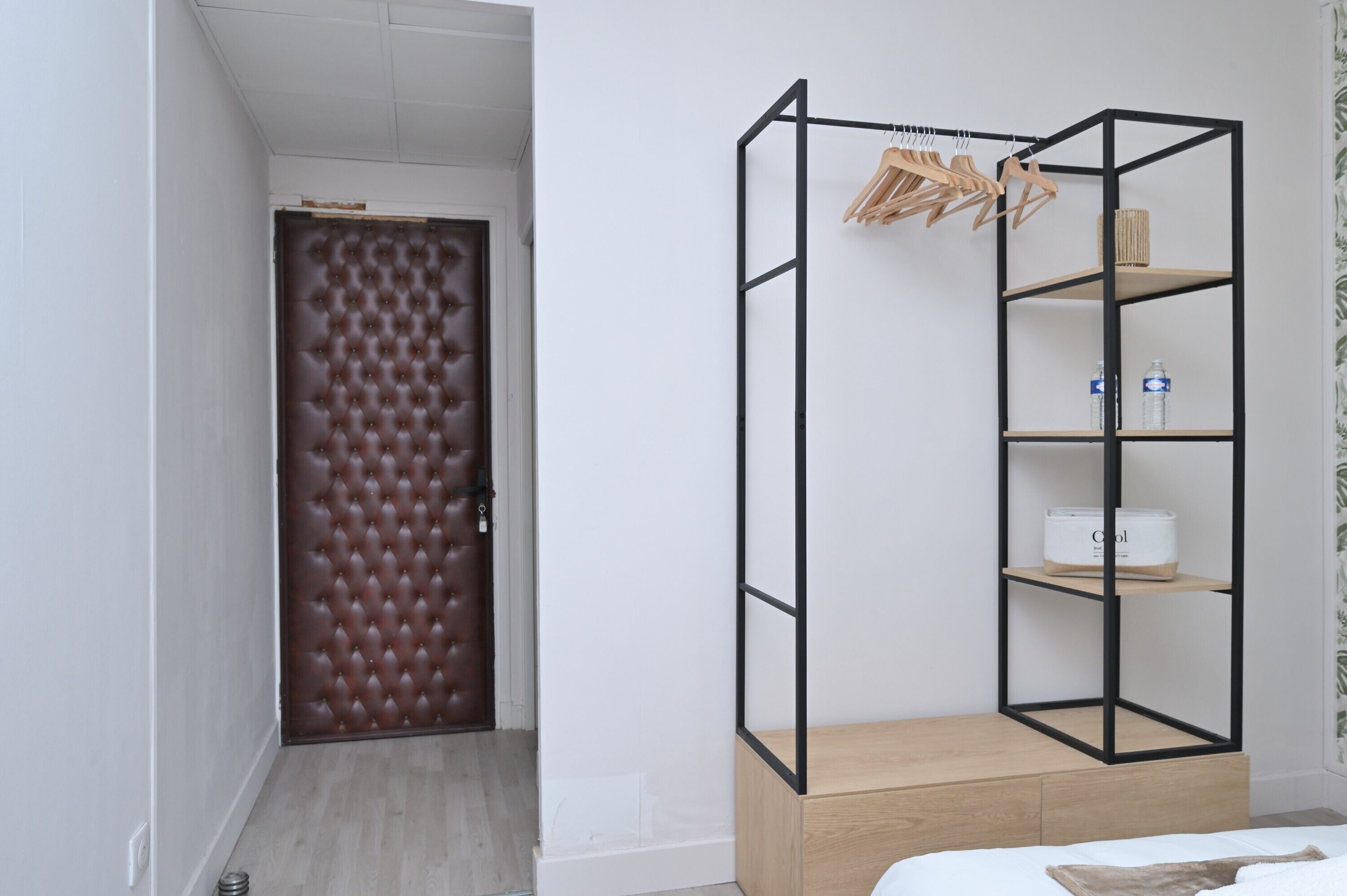 Una habitación con una puerta color café adornada con capitoné, un armario metálico negro con estantes, y una cama con cabecera blanca.