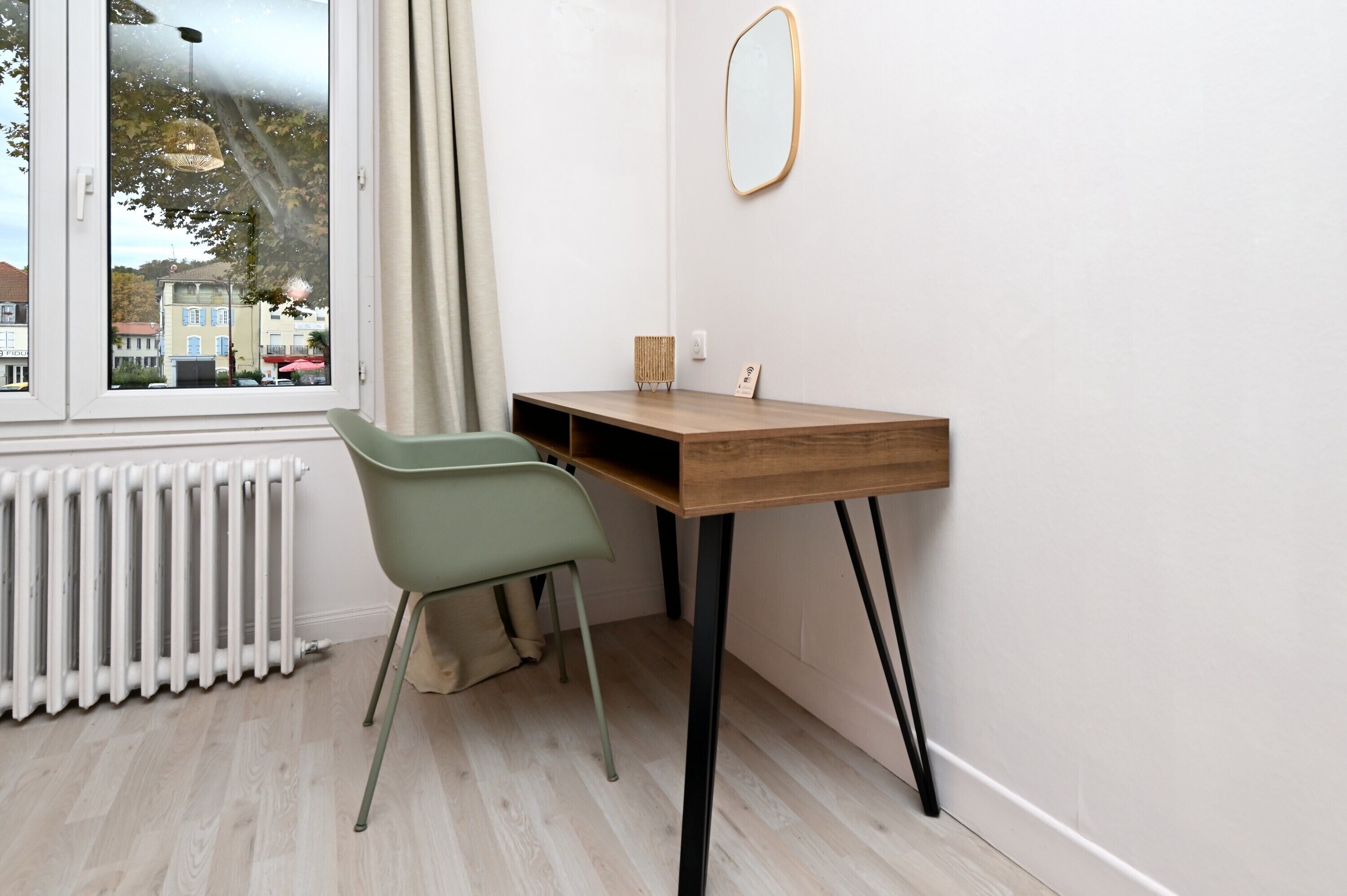 Una stanza con una scrivania in legno, una sedia, un termosifone, una finestra vista edifici e uno specchio fissato alla parete.