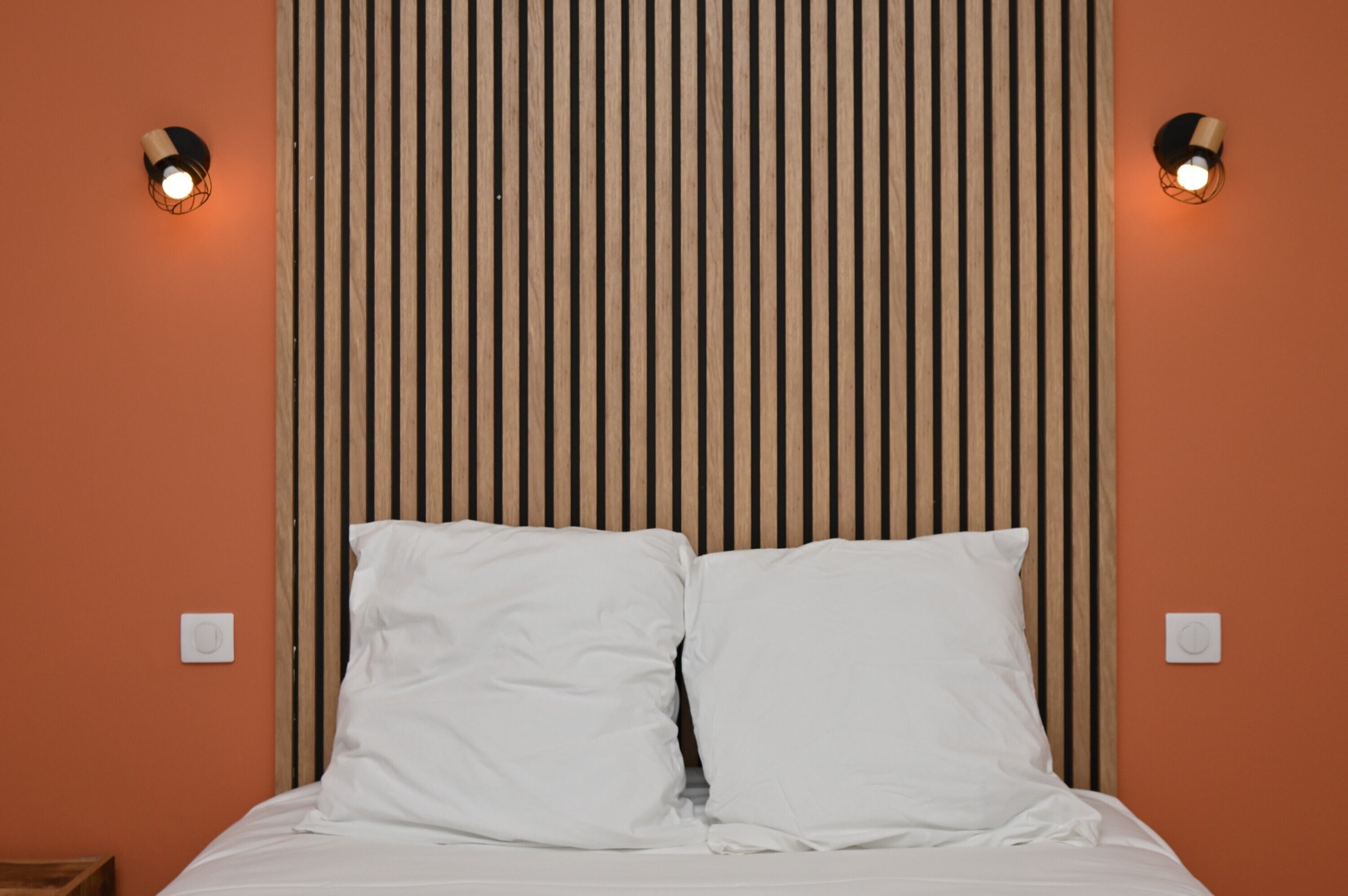 A bed with white bedding, a wooden slat headboard, and two wall-mounted light fixtures.