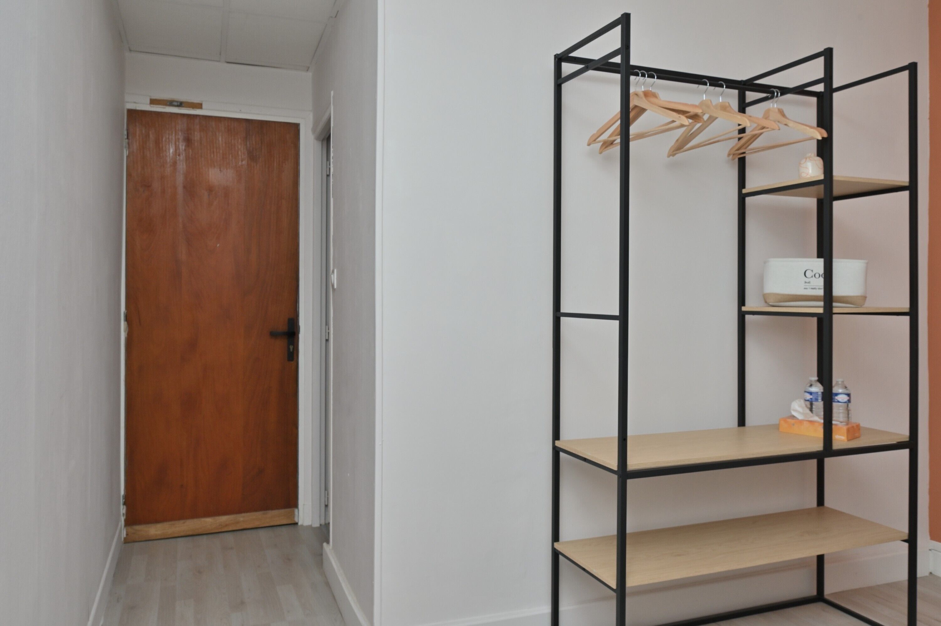 Un couloir avec une porte en bois, une étagère en métal noir avec des cintres en bois, et un récipient blanc portant l’étiquette « Coco ».