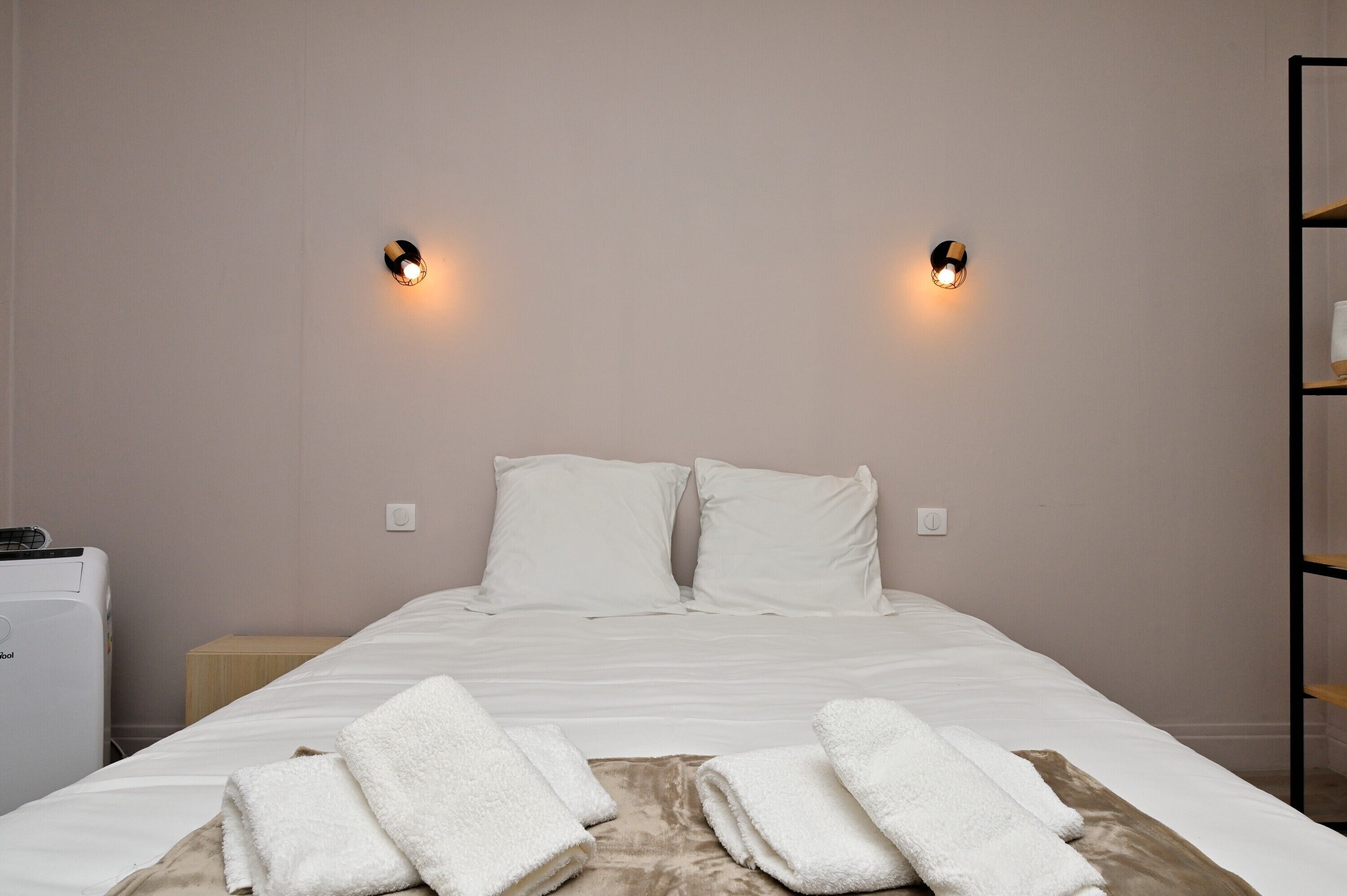 Een netjes opgemaakt bed met witte lakens en twee kussens, een nachtkastje met een lamp, en een wandplank.