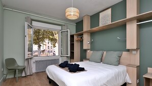 A modern bedroom with a large bed, a window with a view of buildings, a green chair, and a wooden headboard.
