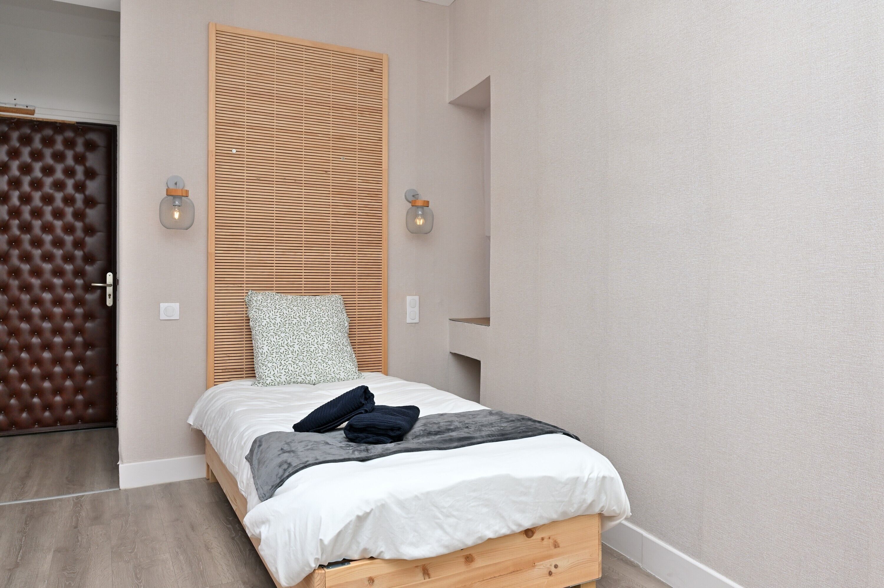 A bedroom with a wooden headboard, a bed with white bedding, two wall-mounted lights, and a door with a patterned panel.