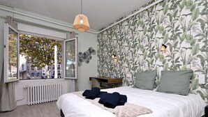 Ein Schlafzimmer mit großem Fenster, Heizkörper, einem Bett mit ordentlich gefalteten Handtüchern und einer Wand mit einem Blattmuster.