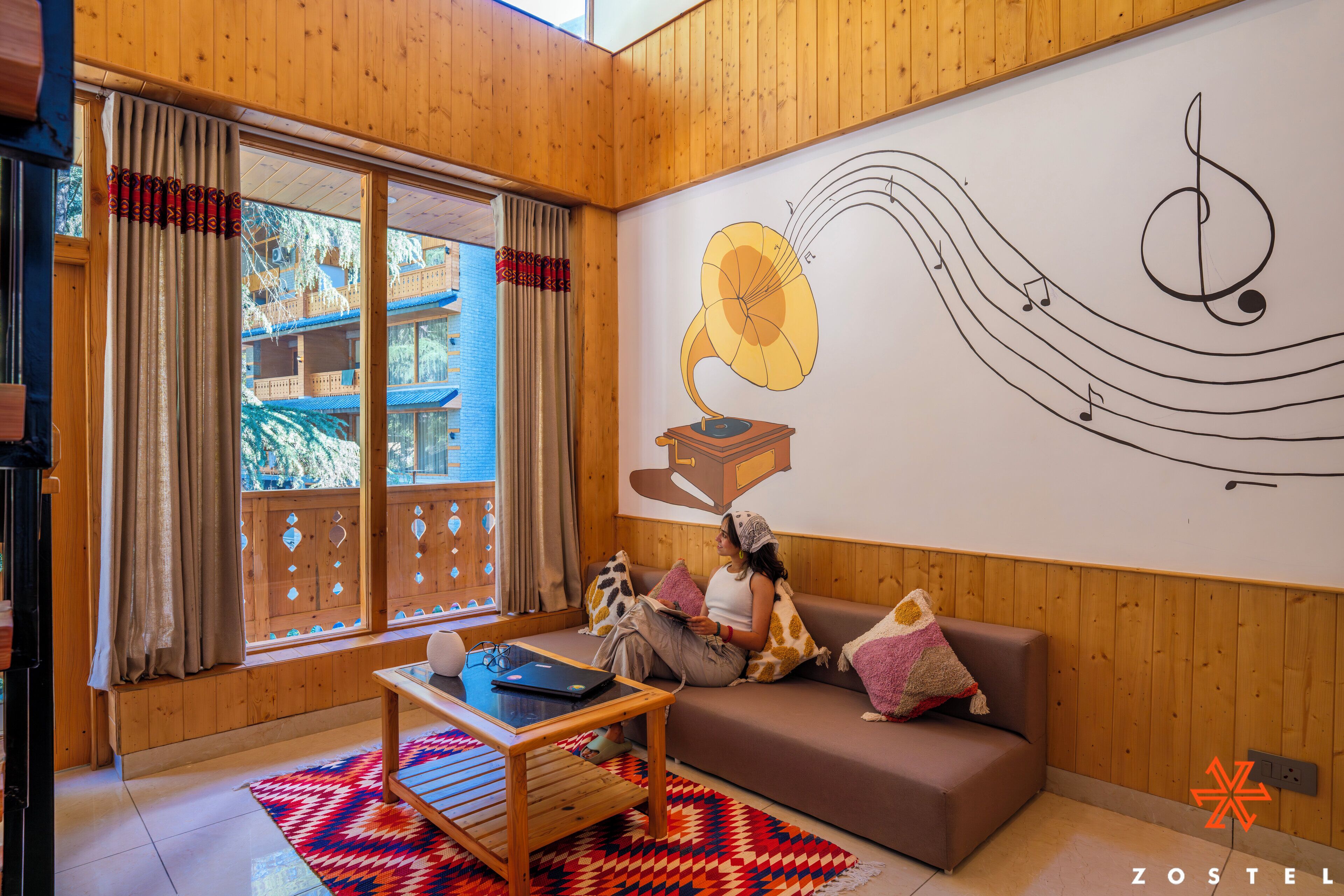 A cozy room with wooden walls, a large window with curtains, a sofa with cushions, a small table, and a wall mural of a gramophone and musical notes.