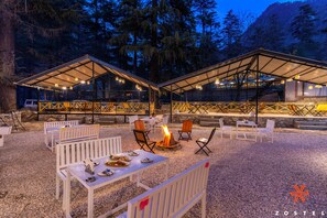 An outdoor dining area with white furniture and a fire pit, surrounded by trees and a mountainous backdrop. - Zostel Kasol (Main Market) (Kasol)