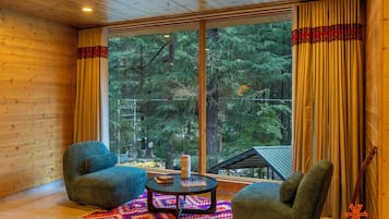 A cozy room with wooden walls, a large window with curtains, two armchairs, a small table with a chessboard, and a guitar leaning against the wall.