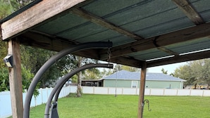 Een hangmat onder een houten pergola in een achtertuin met een gazon, een witte schutting en een huis op de achtergrond.