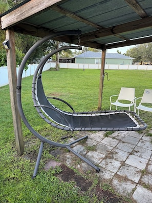 Una hamaca bajo una pérgola de madera en un patio trasero con césped, una cerca blanca y una casa al fondo.