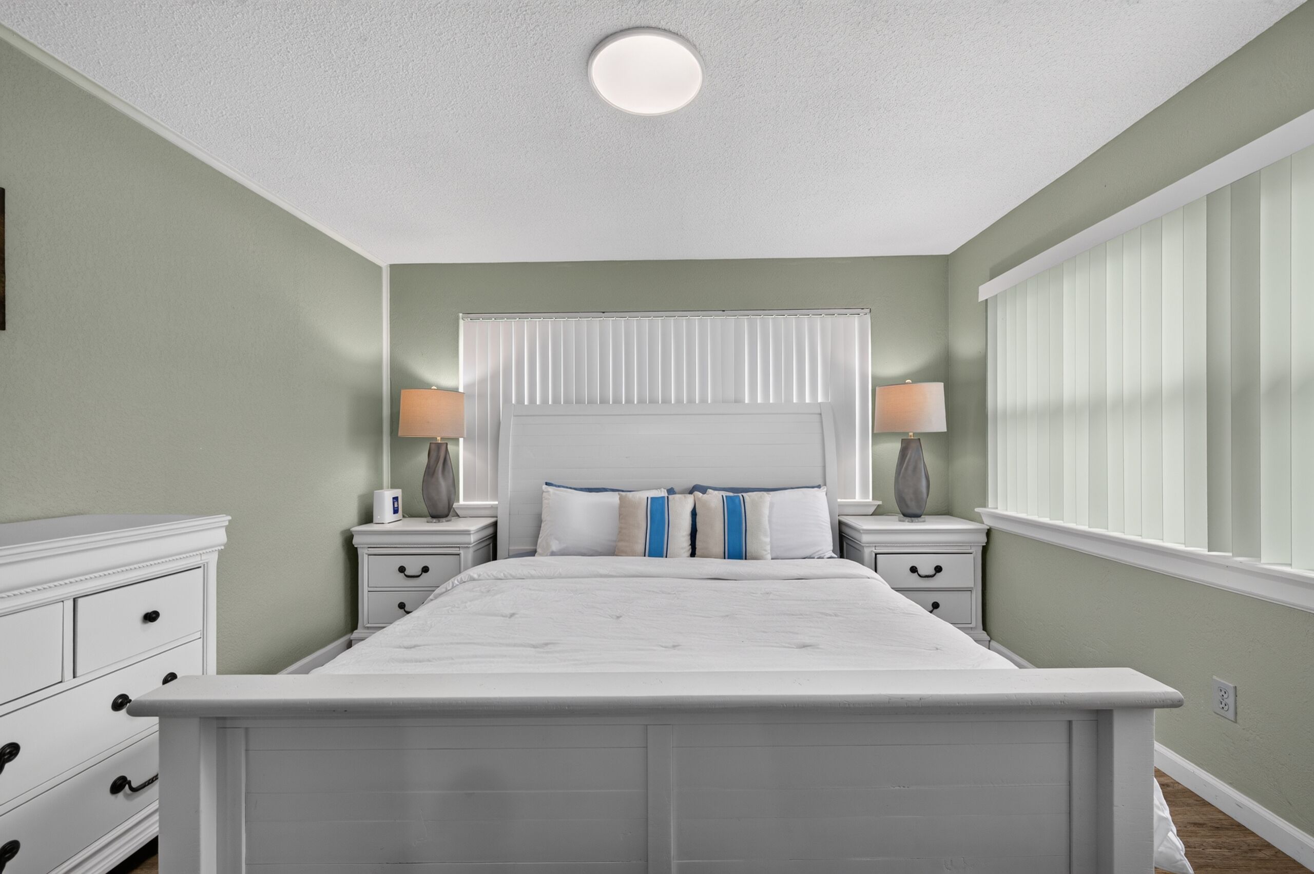 Une chambre à coucher avec un grand lit, deux tables de chevet avec des lampes, une commode et une fenêtre avec des stores.