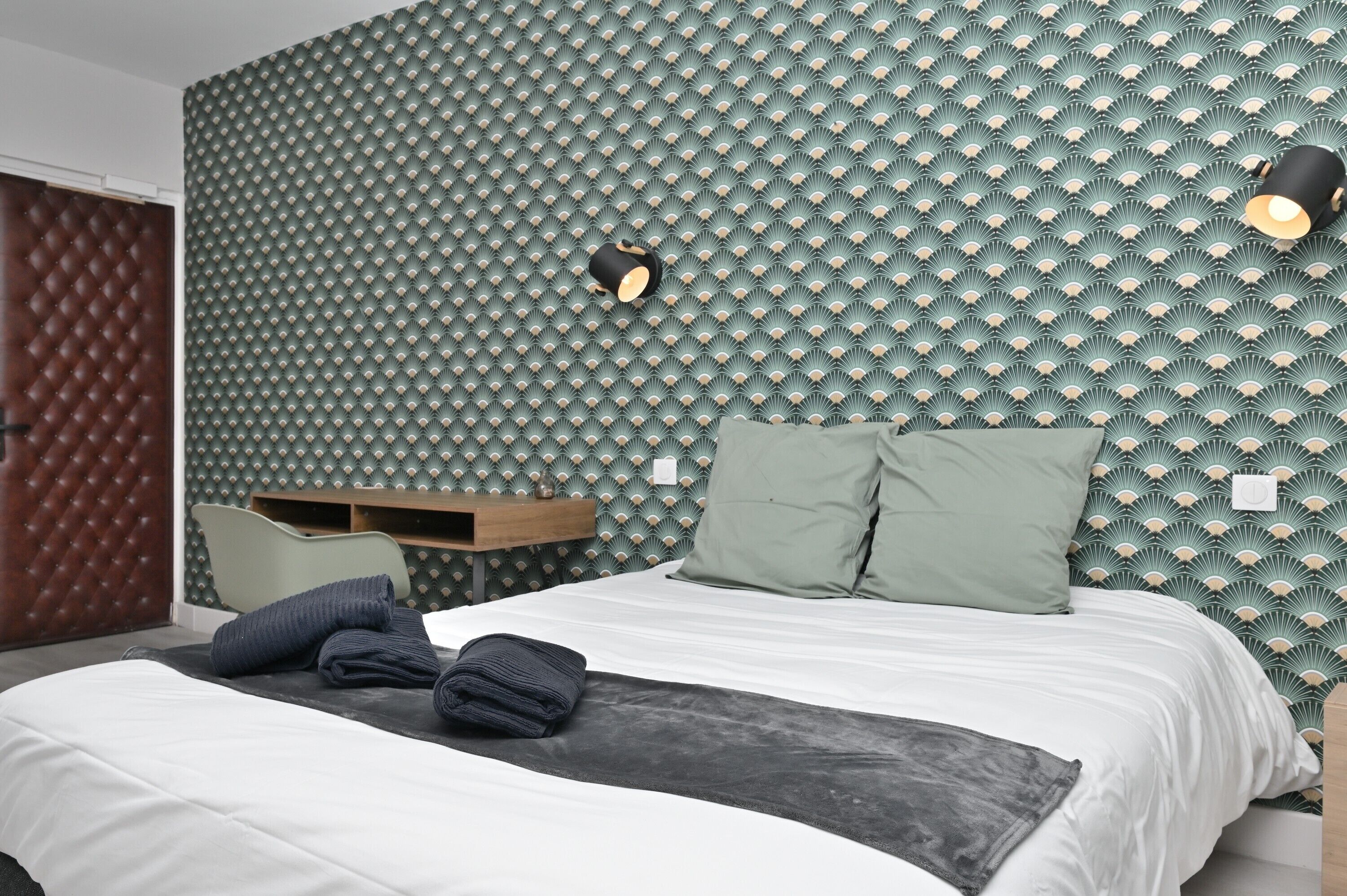 Une chambre d’hôtel moderne avec un mur orné d’un motif, un lit avec des serviettes soigneusement pliées, une table de chevet en bois et deux lampes de lecture fixées au mur.