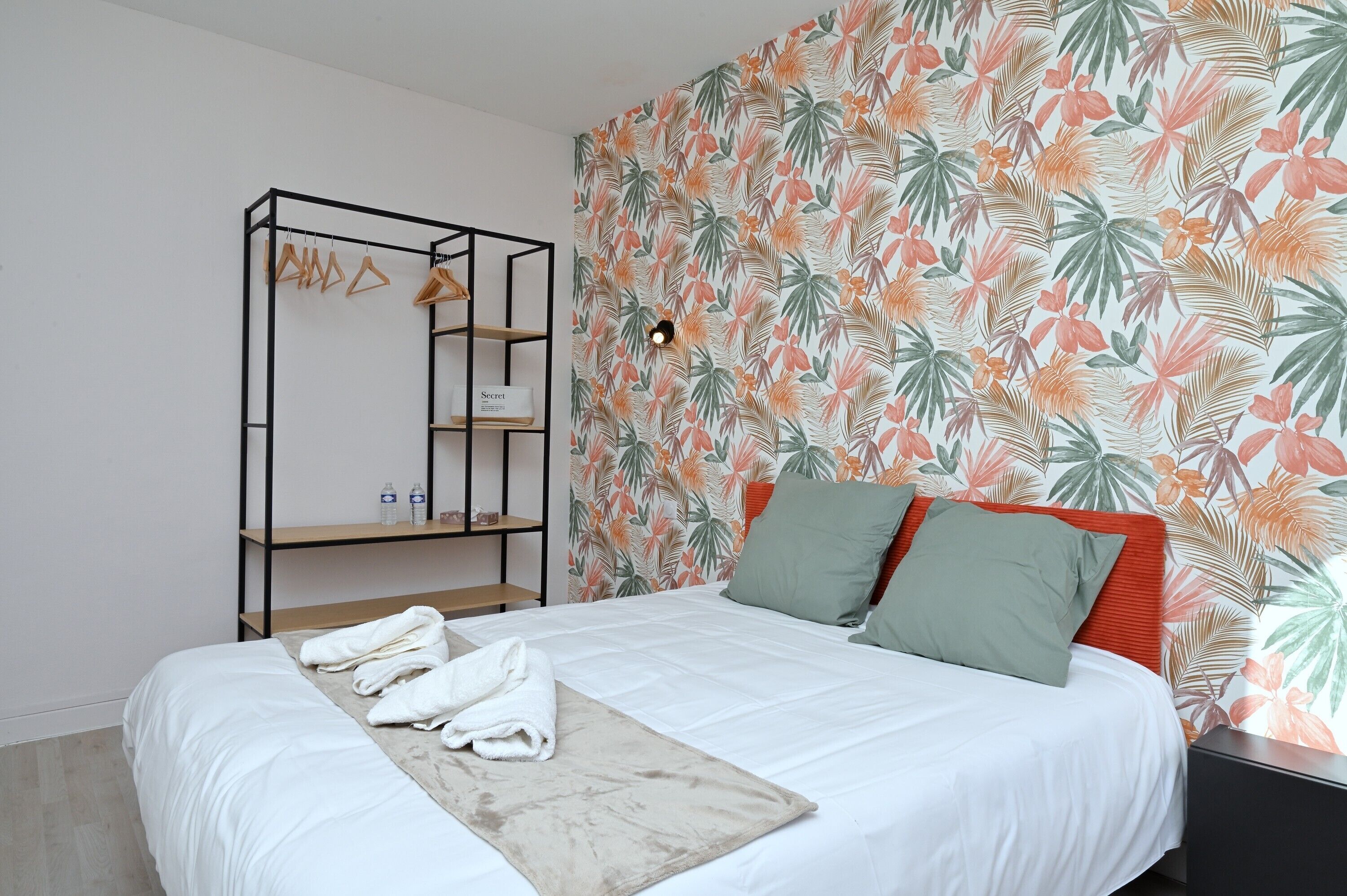 Un dormitorio con cama, una estantería de madera y un papel tapiz con motivos de hojas tropicales.