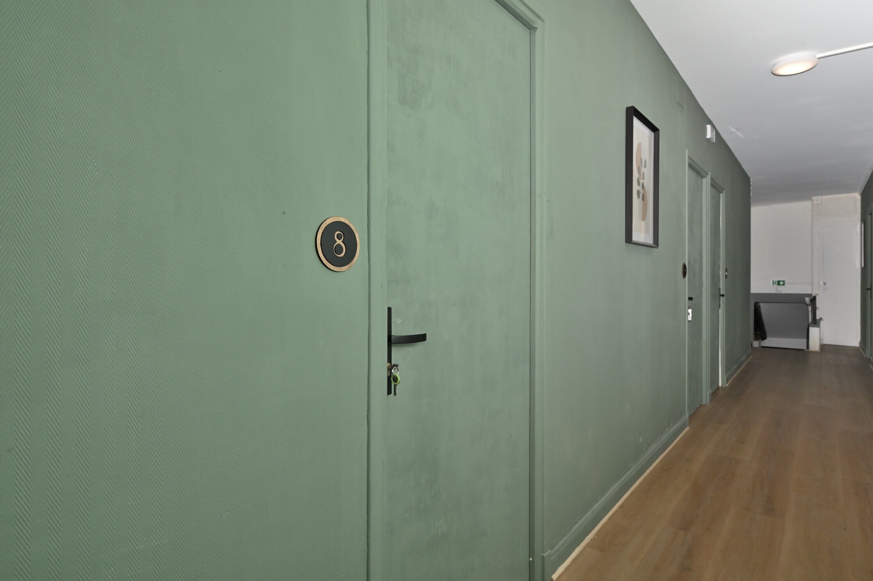 Un couloir aux murs verts, avec un plancher en bois et des portes portant des numéros.
