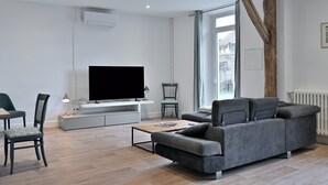 Een moderne woonkamer met een flatscreen televisie, een grijze bank, houten balken en een groot raam.