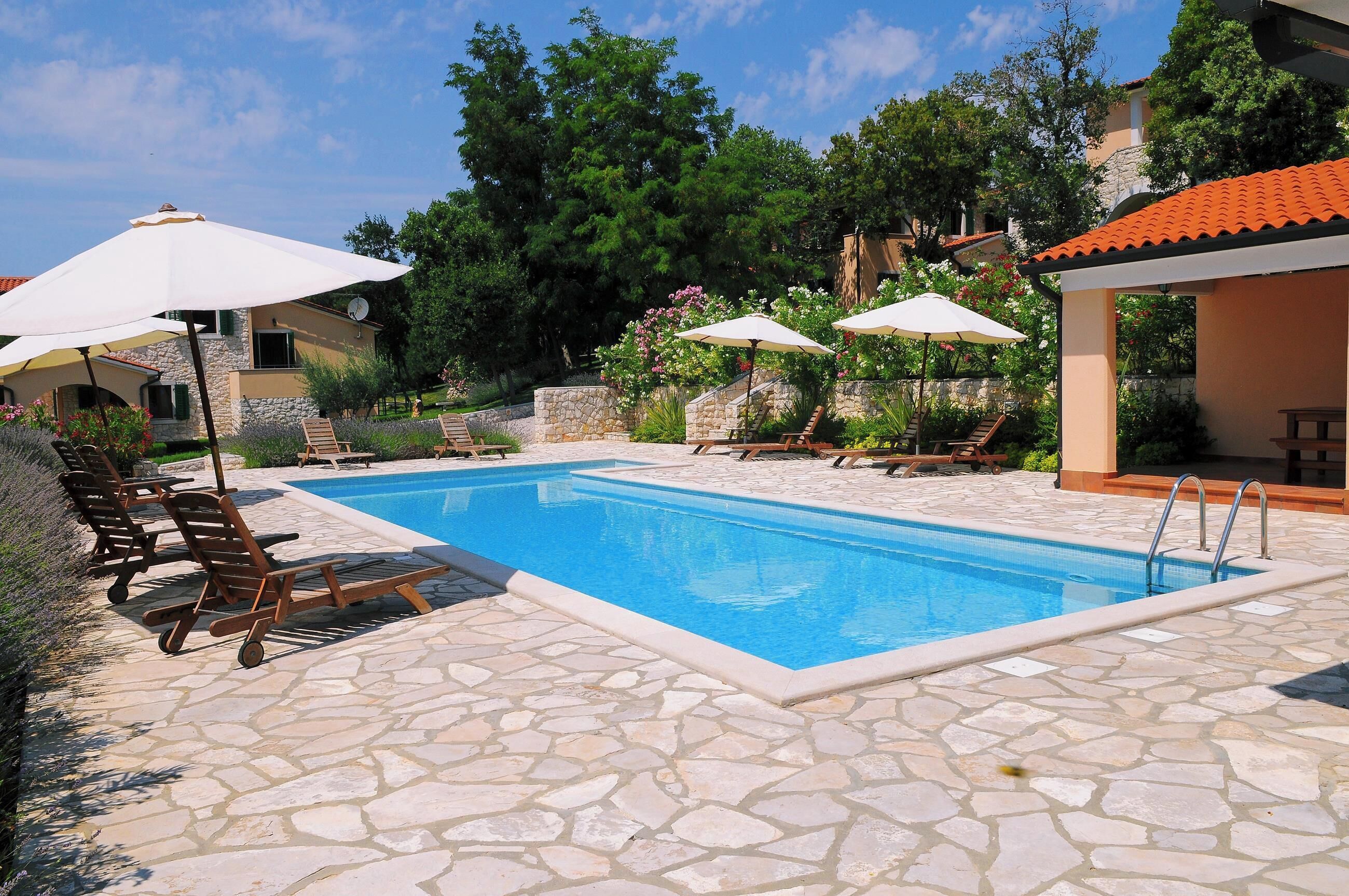 Ein Pool mit Liegestühlen und Sonnenschirmen, umgeben von Steinböden und Grünpflanzen.