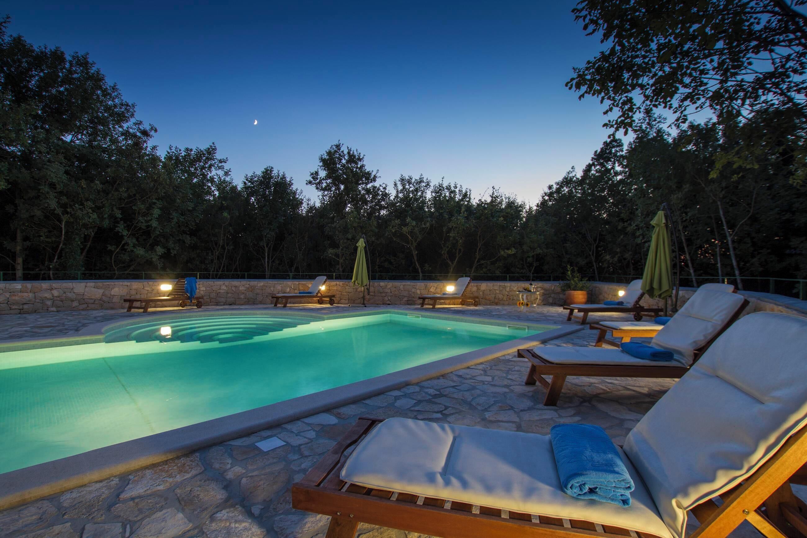 Piscina con sillas de descanso y sombrillas, rodeada de muros de piedra y árboles.