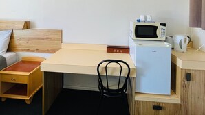 Ein kleines Hotelzimmer mit Bett, einem kleinen Holztisch, einem Stuhl sowie einer Küchenzeile mit Mikrowelle und Wasserkocher. Ein Schild zeigt das Wort 'TEA'.
