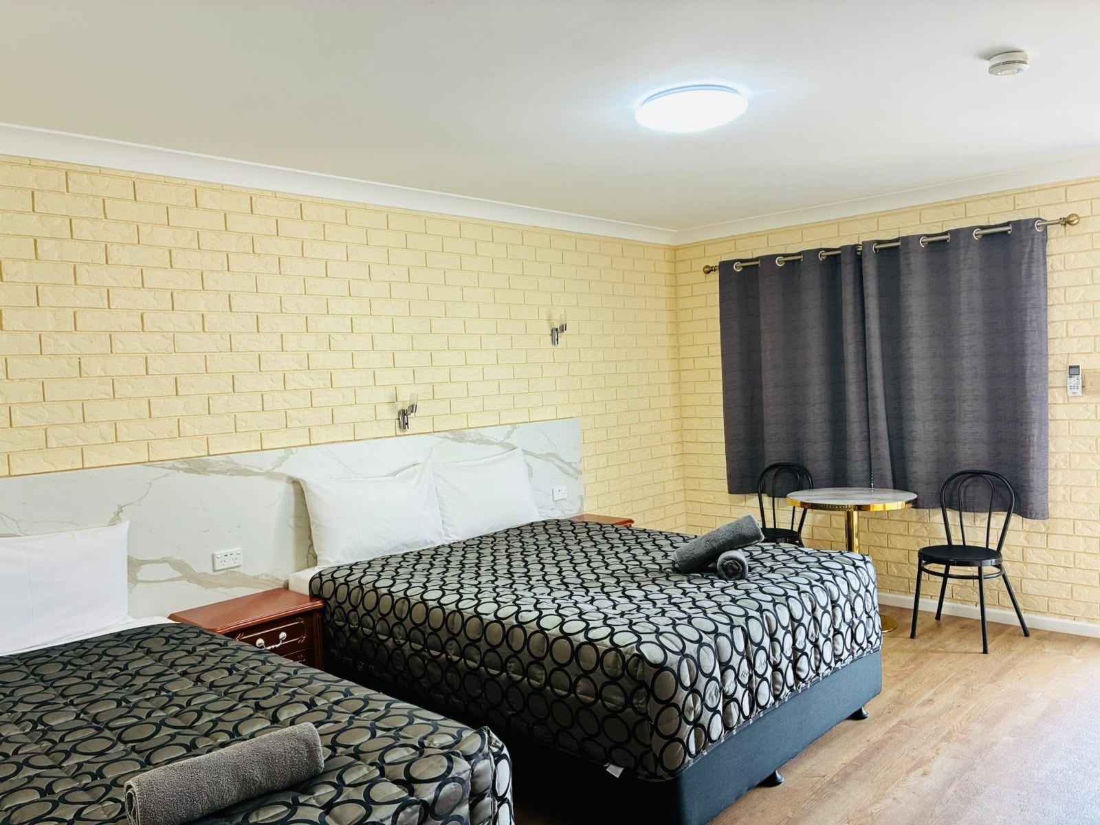 Een hotelkamer met twee bedden, een tafeltje, twee stoelen, een muur met baksteenmotief en een gordijn.
