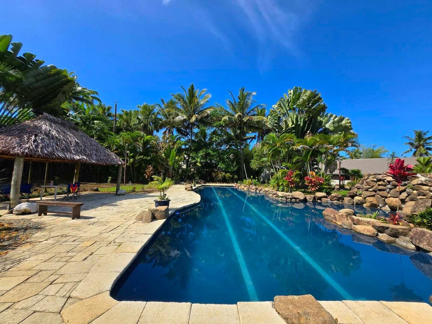 Uma piscina com borda de pedra, cercada por vegetação tropical e uma estrutura coberta de palha.