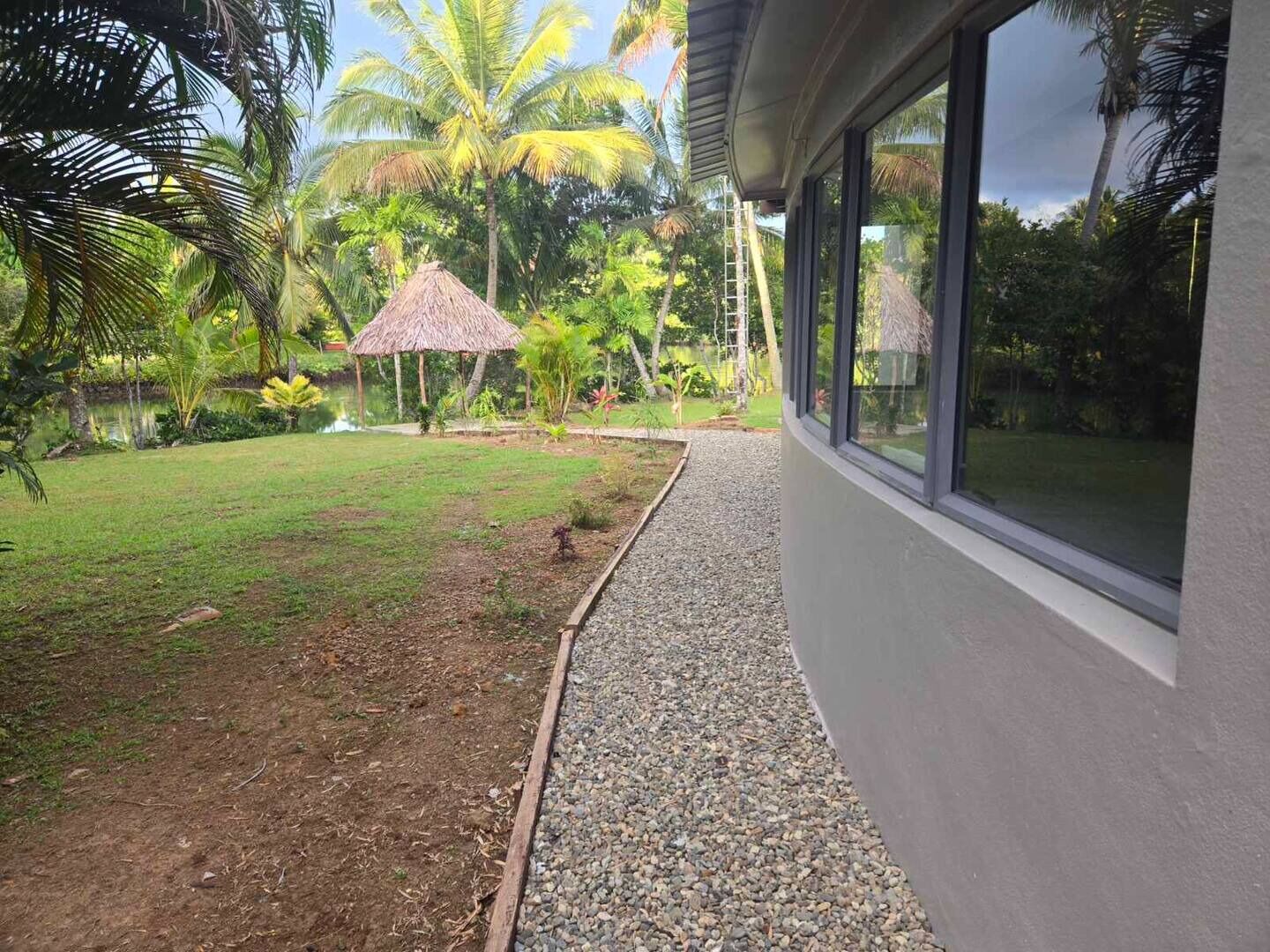 Um edifício moderno com grandes janelas que dão para um jardim bem cuidado, com uma construção de telhado de palha, palmeiras e um caminho de cascalho.