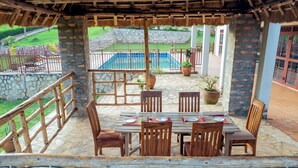 Une terrasse couverte avec un sol en pierre, des tables et des chaises en bois, et une vue sur une piscine et un jardin.