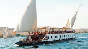 A large boat with multiple decks and white sails, likely used for tours or transportation, on a body of water with a desert landscape in the background. - Sonesta Dahabeya Amirat I - Nile Cruise Ship - 7 nights from Luxor every Saturday  (Luxor)