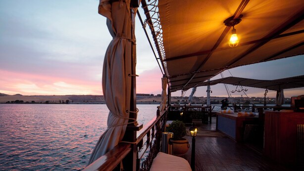 A deck with a view of the water, a wooden railing, a cushioned bench, and a hanging light fixture. - Sonesta Dahabeya Amirat I - Nile Cruise Ship - 7 nights from Luxor every Saturday  (Luxor)