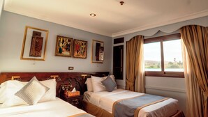 A hotel room with two beds, a large window with curtains, and several framed artworks on the wall. - Sonesta Dahabeya Amirat I - Nile Cruise Ship - 7 nights from Luxor every Saturday  (Luxor)