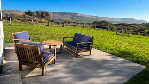 A patio with two wooden chairs and a small round table, overlooking a grassy field and distant hills.