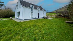 A white two-story house with a dark roof, a front porch, and a small garden area. - Ballylusky Holiday Home (Ballydavid)