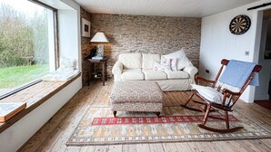 A cozy living room with a stone wall, a white sofa, a patterned ottoman, a wooden rocking chair, a lamp, and a dartboard. - Ballylusky Holiday Home (Ballydavid)