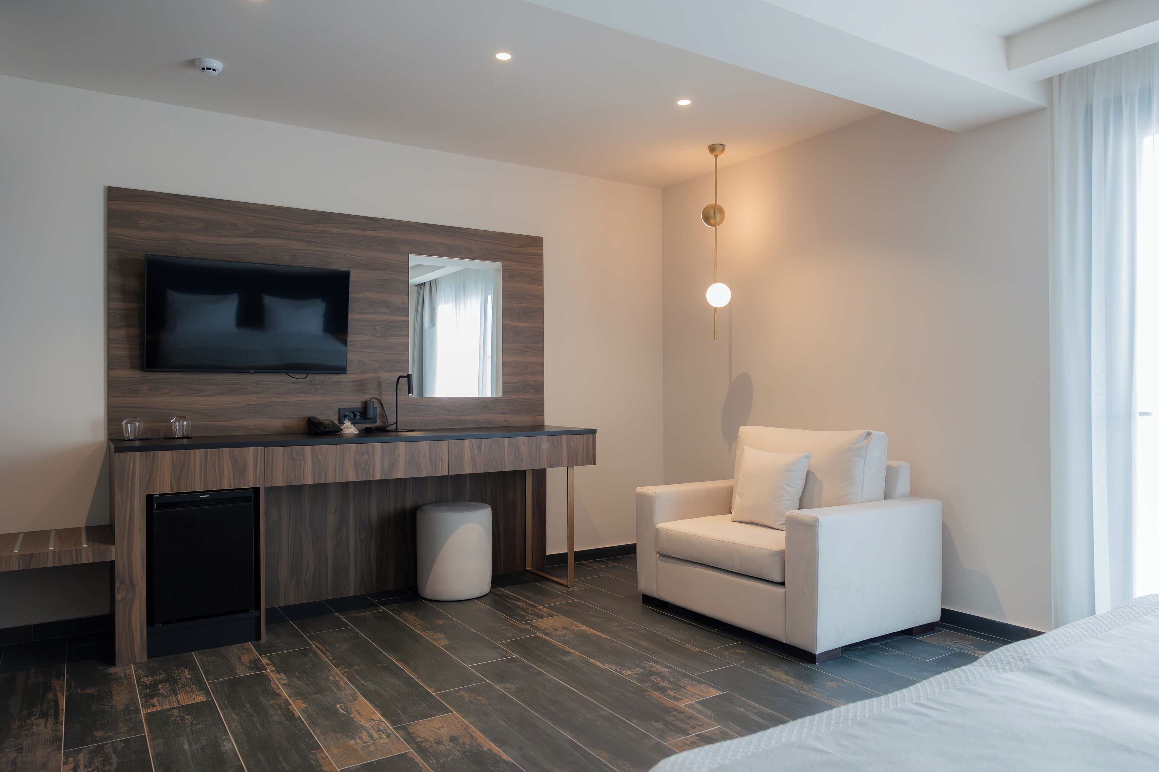 A modern hotel room with a wooden vanity, a flat-screen TV, a white armchair, and a large bed.