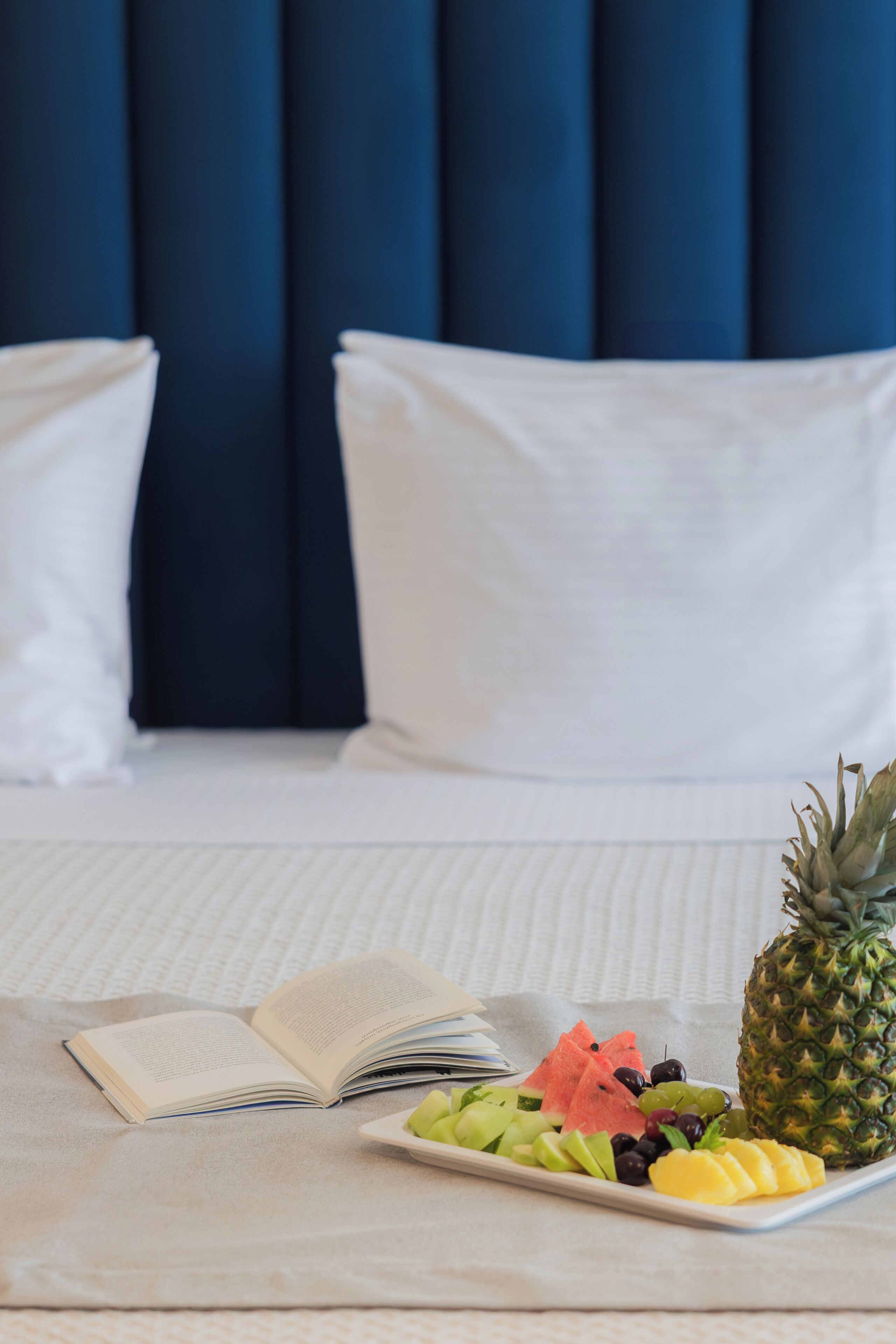 A neatly made bed with a white and blue color scheme, an open book, and a fruit platter on the bed.