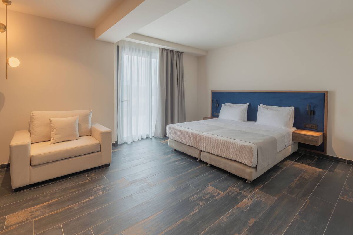 A modern hotel room with a bed, a sofa, a nightstand, and a large window with curtains.