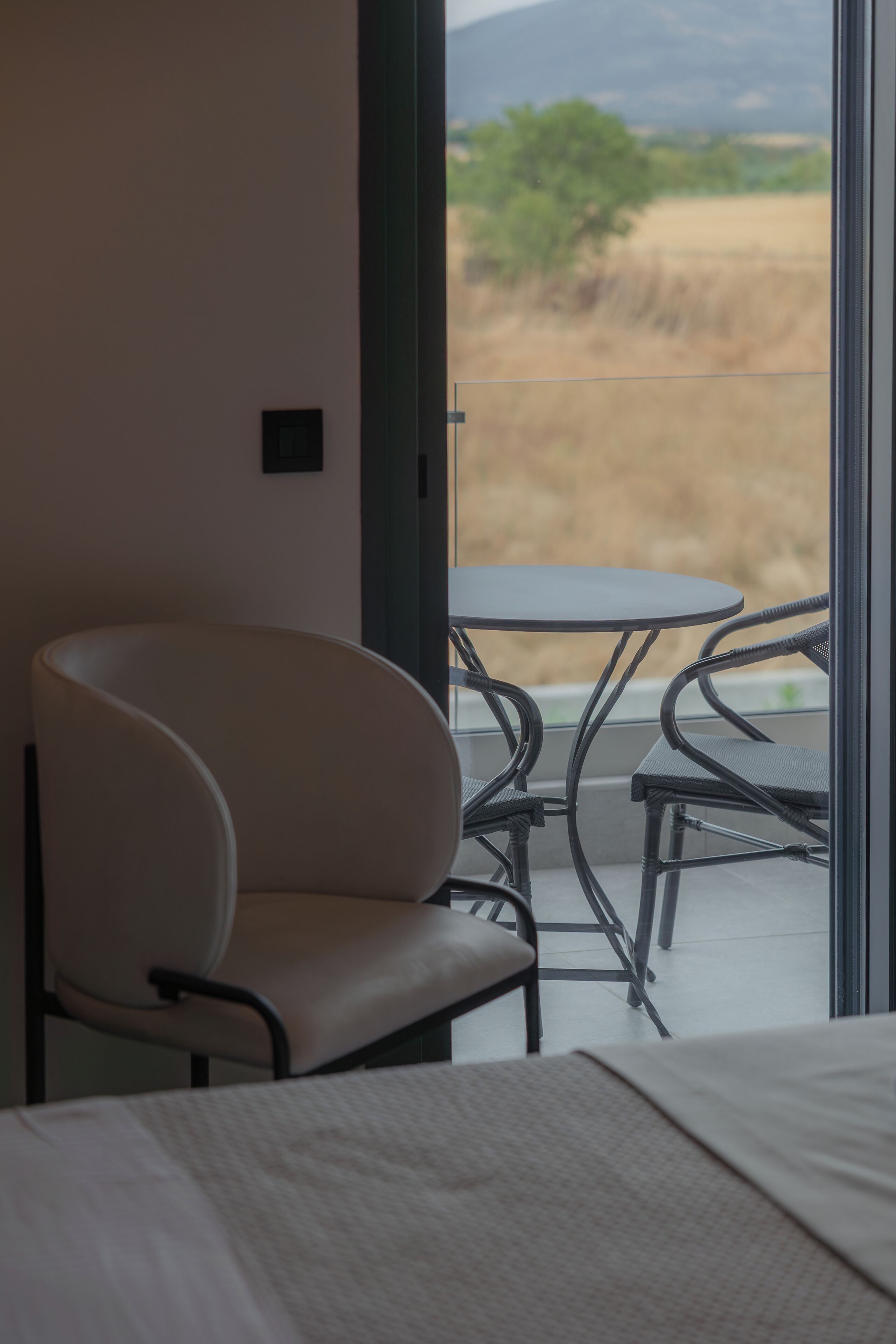 A room with a bed, a chair, and a table with a view of a field and mountains.
