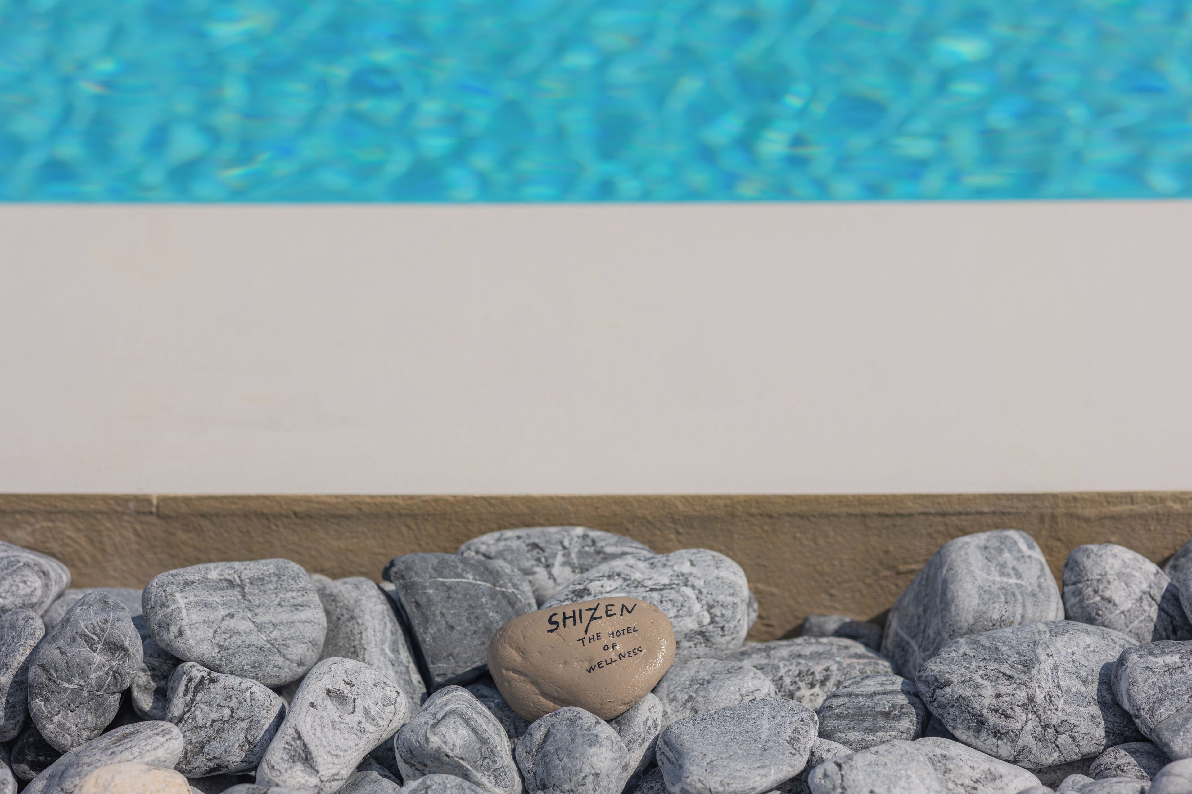 A stone with "SHIZEN THE HOTEL OF WELLNESS" engraved, surrounded by smooth stones, with a pool in the background.