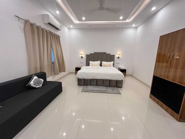 A modern bedroom with a large bed, a black sofa, a wooden wardrobe, and a ceiling fan.