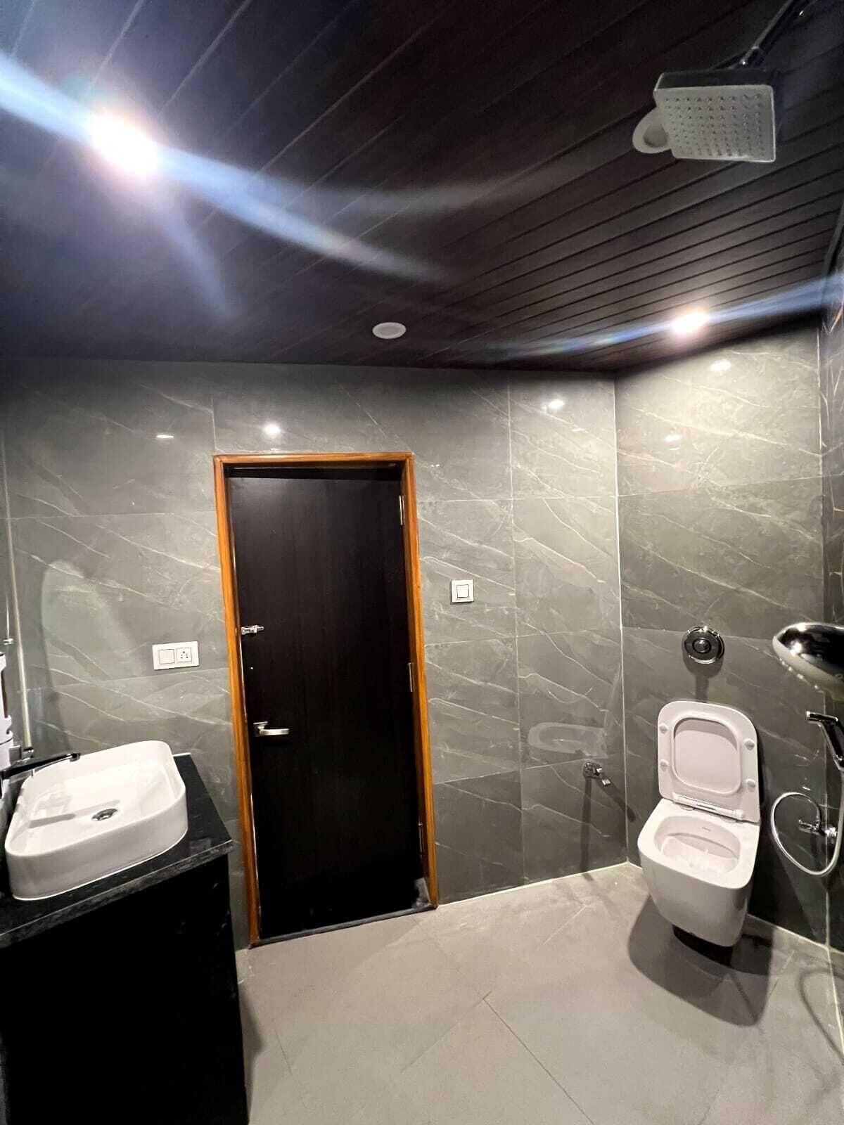 Un baño moderno con pared de mármol, puerta de madera, lavamanos blanco y un inodoro con cabezal de ducha.