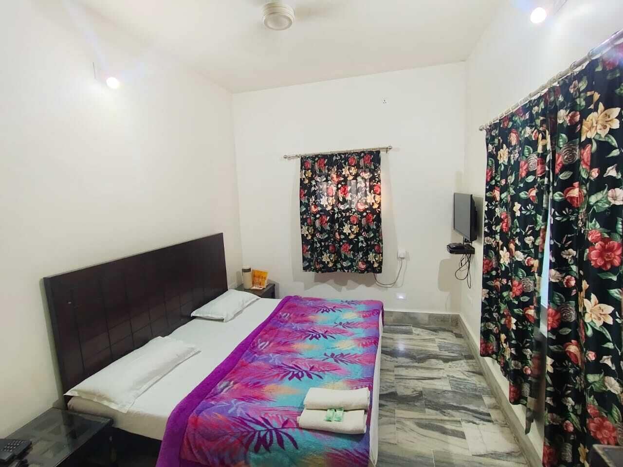 A bedroom with a bed, a nightstand, a wall-mounted TV, and a window with floral curtains.