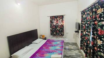 Quarto com cama, mesa de cabeceira, televisão fixada na parede e janela com cortinas florais.