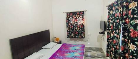 Un dormitorio con cama, mesita de noche, televisor de pared y ventana con cortinas florales.