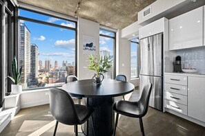Uma cozinha moderna com uma mesa de jantar redonda, cadeiras pretas, uma planta em vaso e vista para a cidade através de amplas janelas.