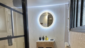 A modern bathroom with a wooden vanity, a round mirror with LED lighting, and a small table with a box on it. - Gîte PREY DES VOSGES (Prey)