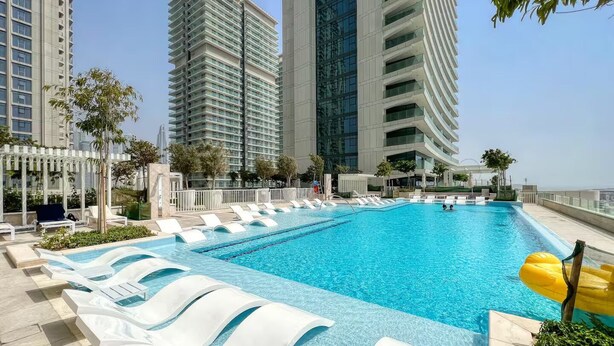 A rooftop swimming pool with white lounge chairs, surrounded by tall buildings and greenery. - Beach Vista Residences - Dubai Harbour (Dubai)